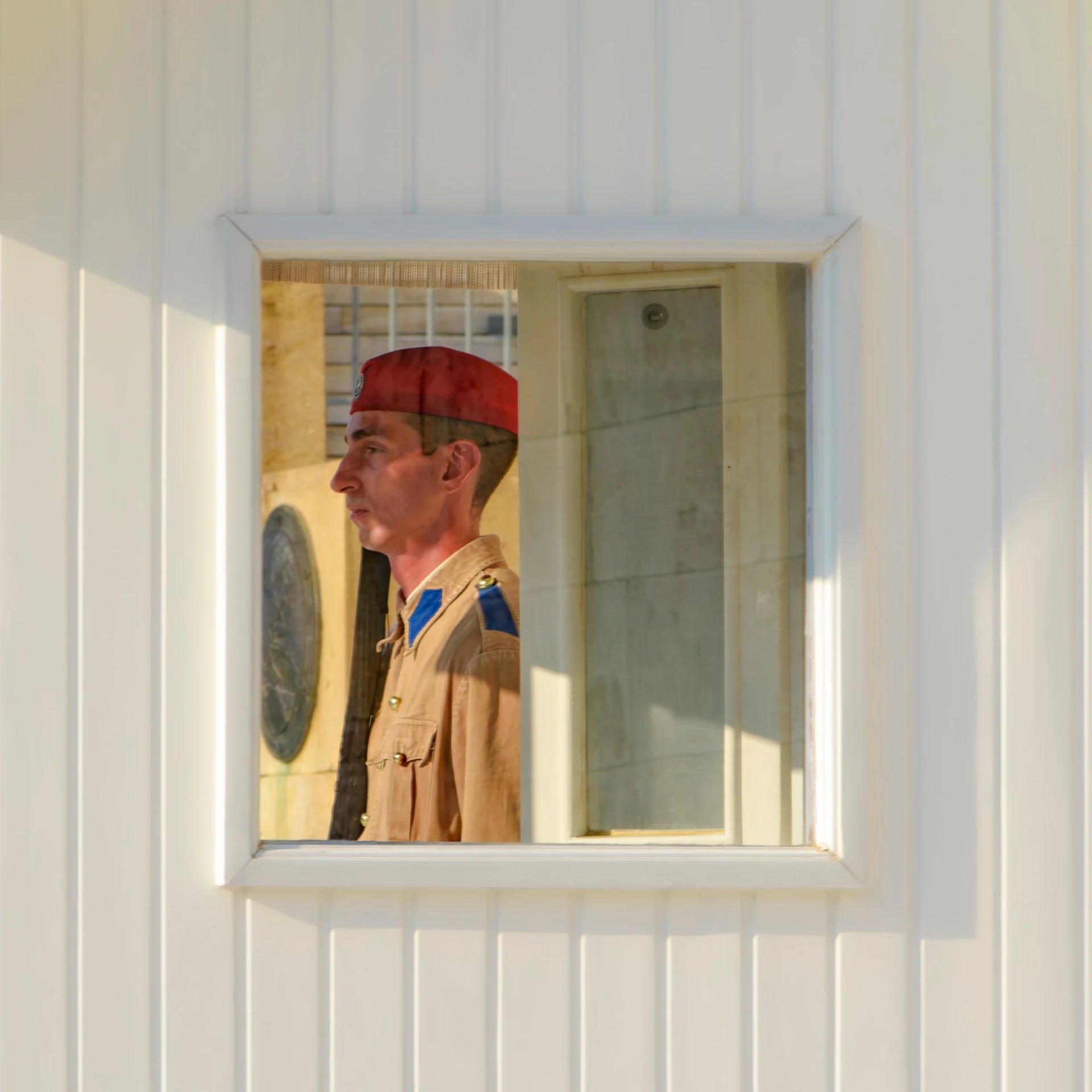 A guard in tan is framed in profile by the small square window of a white sentry box.