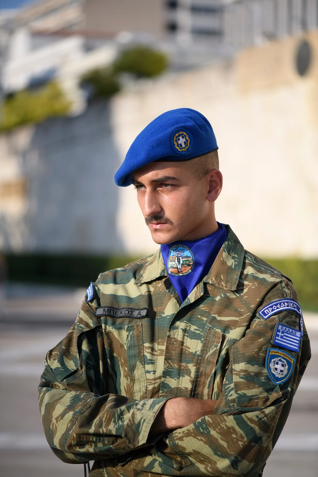 A guard in camouflage and a blue beret stands with folded arms against a softly blurred background.