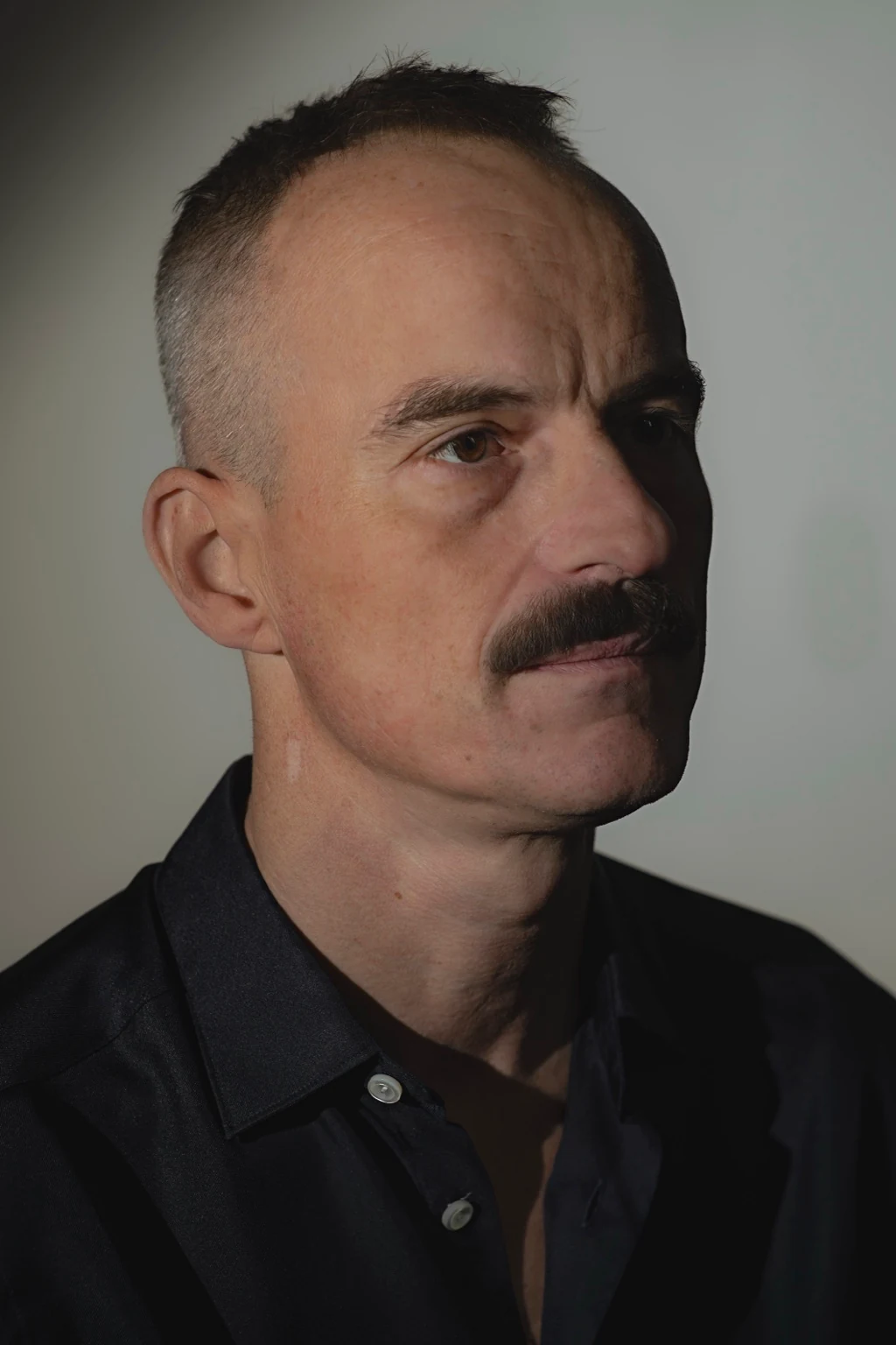 A side-lit close portrait of a moustached sitter in a dark shirt.