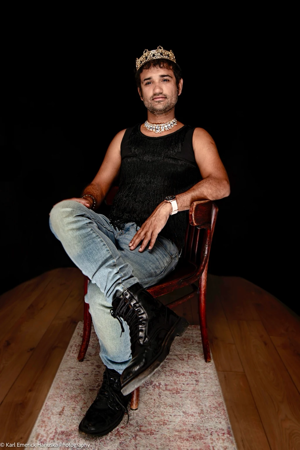 A sitter in an embroidered cap, black top, jeans and boots sits cross-legged on a wooden chair.