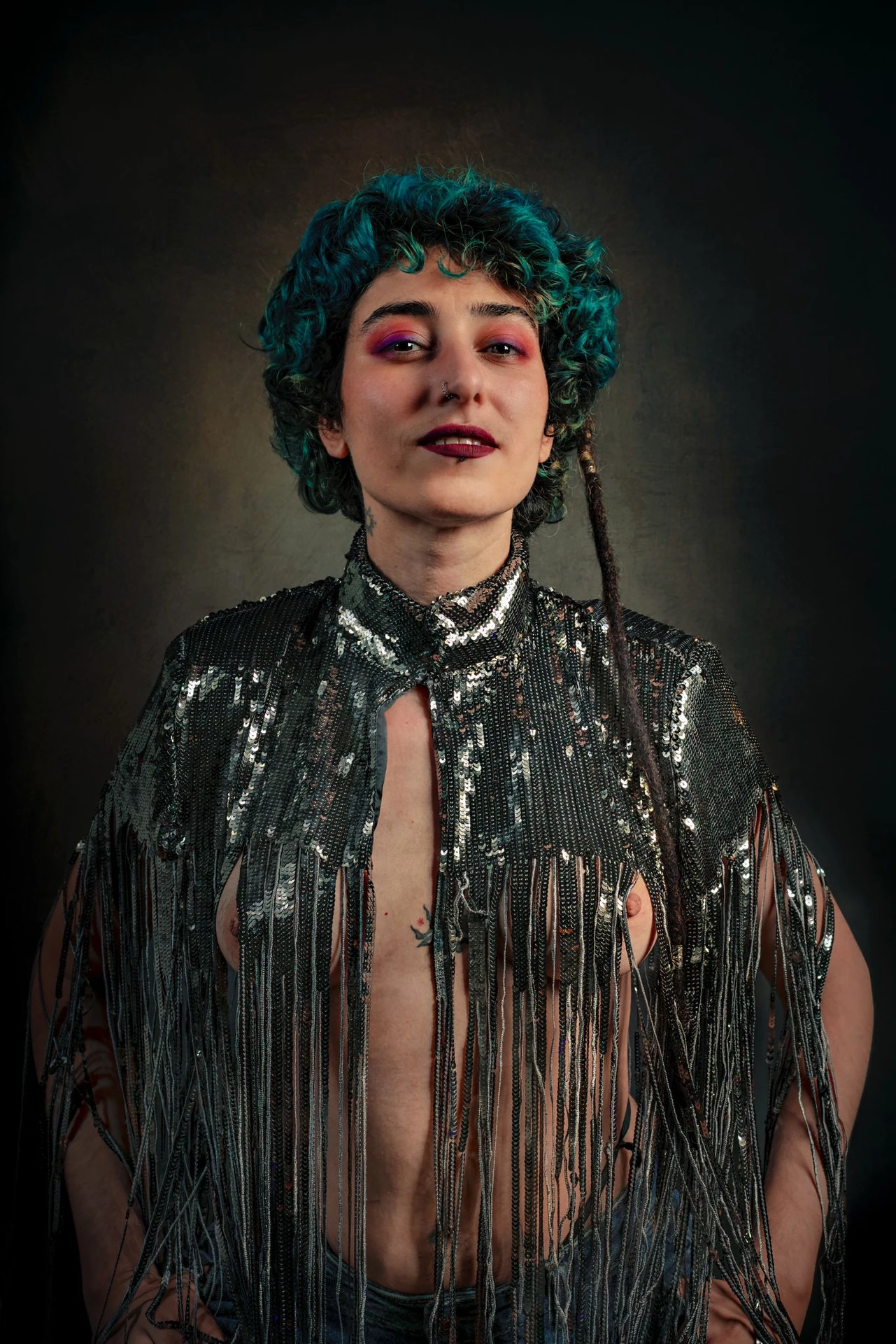 A sitter with teal curls and colourful eye make-up wears a silver fringed top against a dark backdrop.