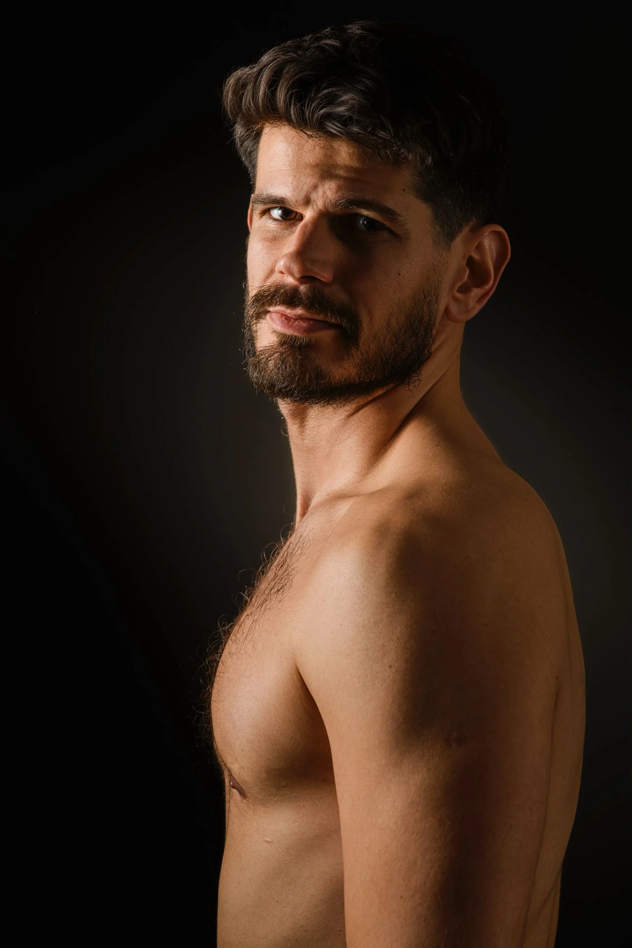 A bare-chested sitter turns toward the camera, with side light tracing the shoulder and face against darkness.