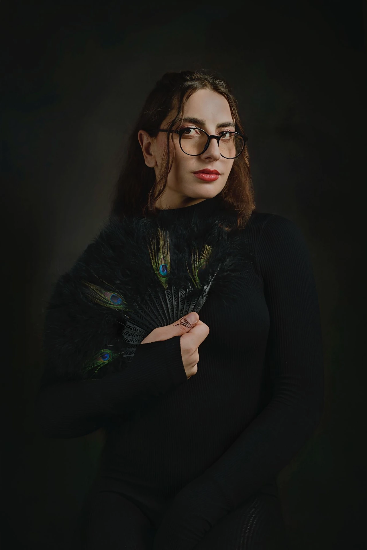A sitter in glasses and a black high-necked outfit holds a dark feather fan against the chest.