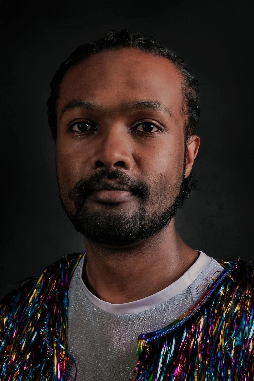 A close portrait with a direct gaze and an iridescent fringed jacket against a dark background.