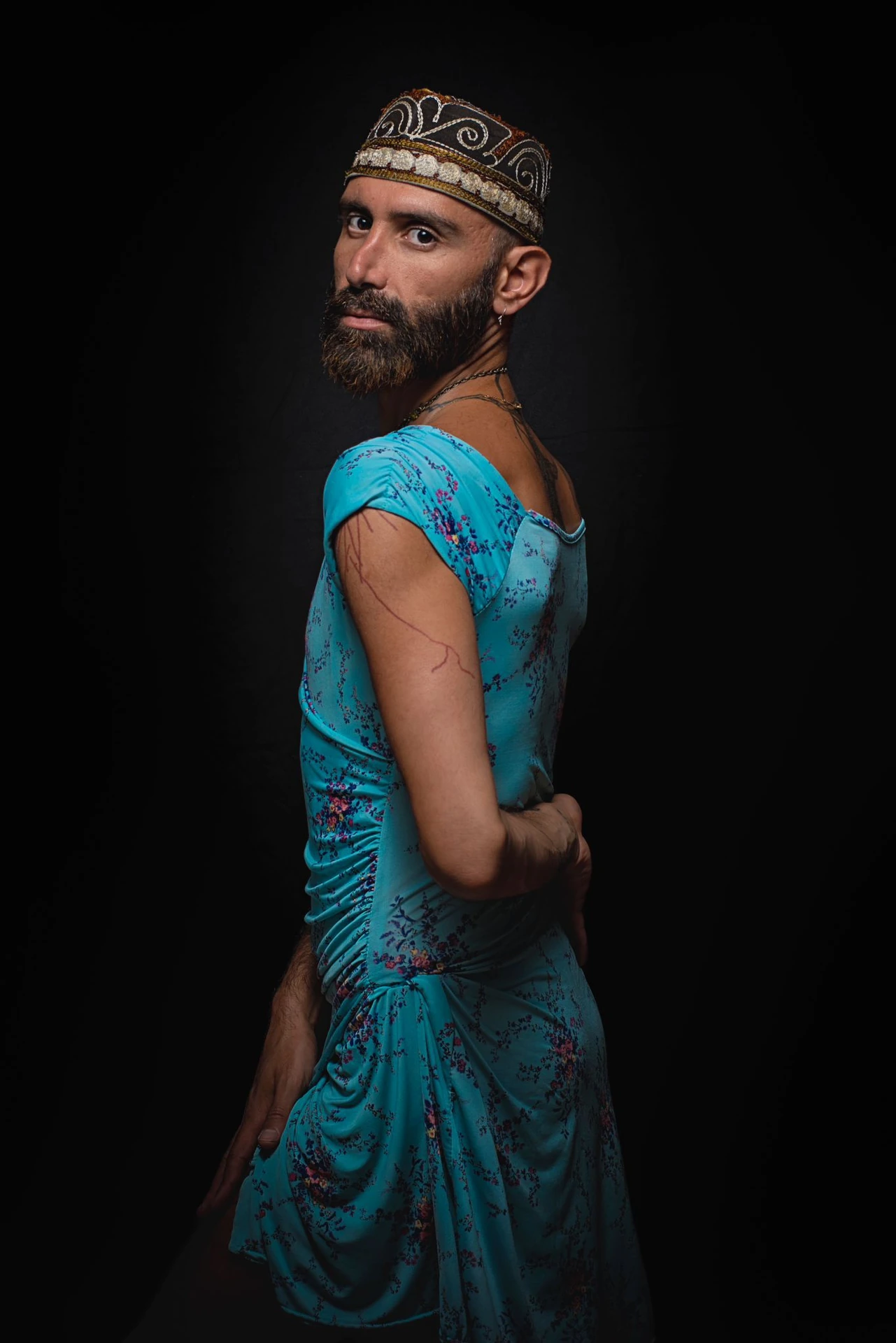 A bearded sitter in an embroidered cap and blue floral dress turns back toward the camera.