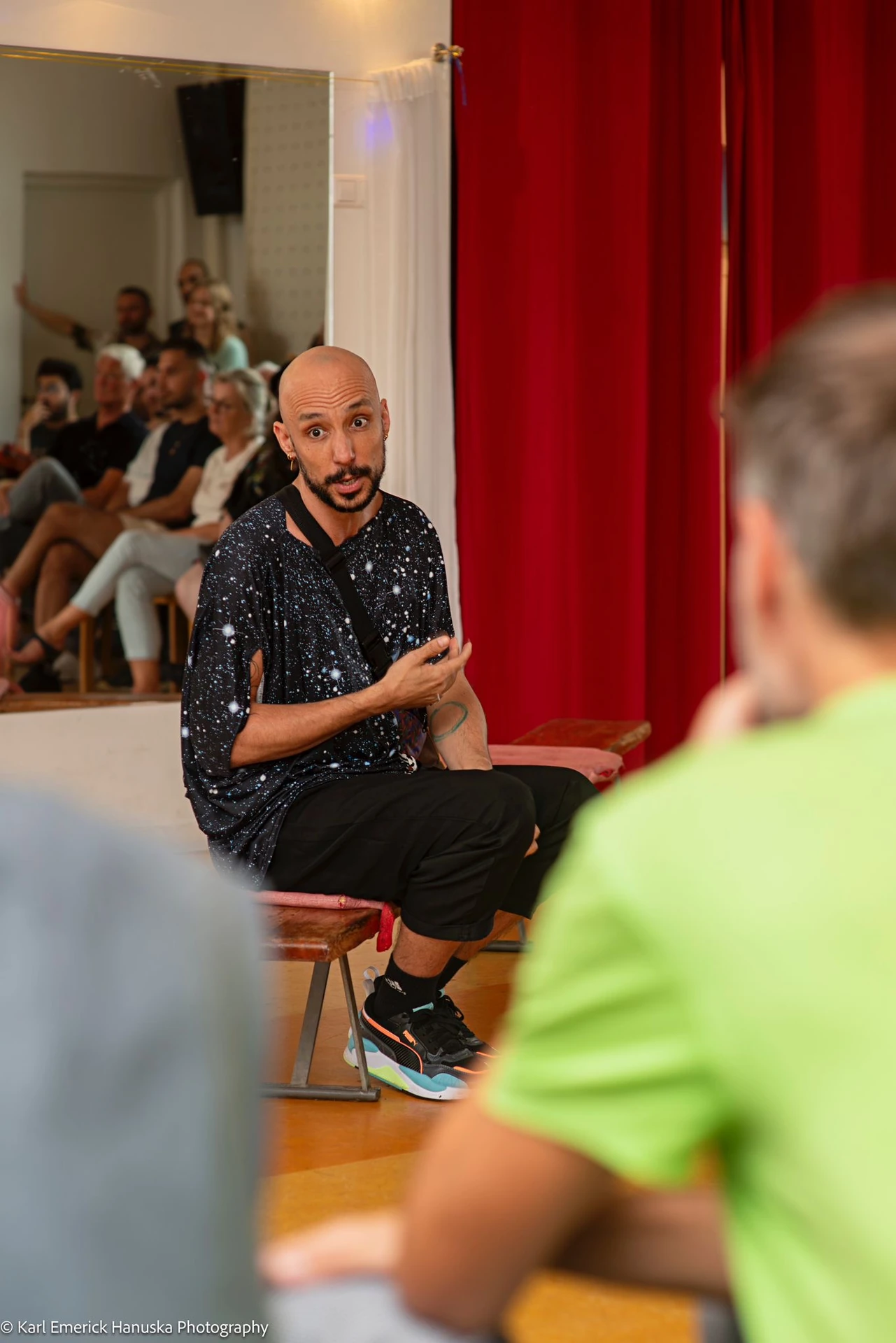 A seated participant gestures during a conversation beside a red curtain, with listeners visible in the mirror.