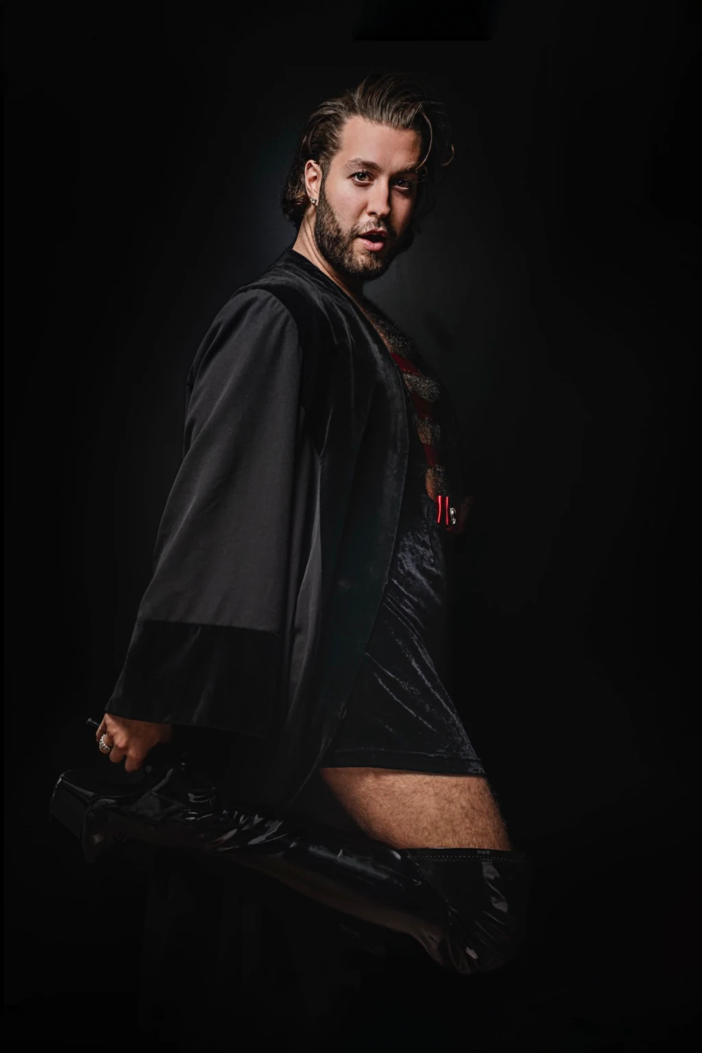 Nojan wears a long black cape and red harness, holding the heel of a raised black boot.