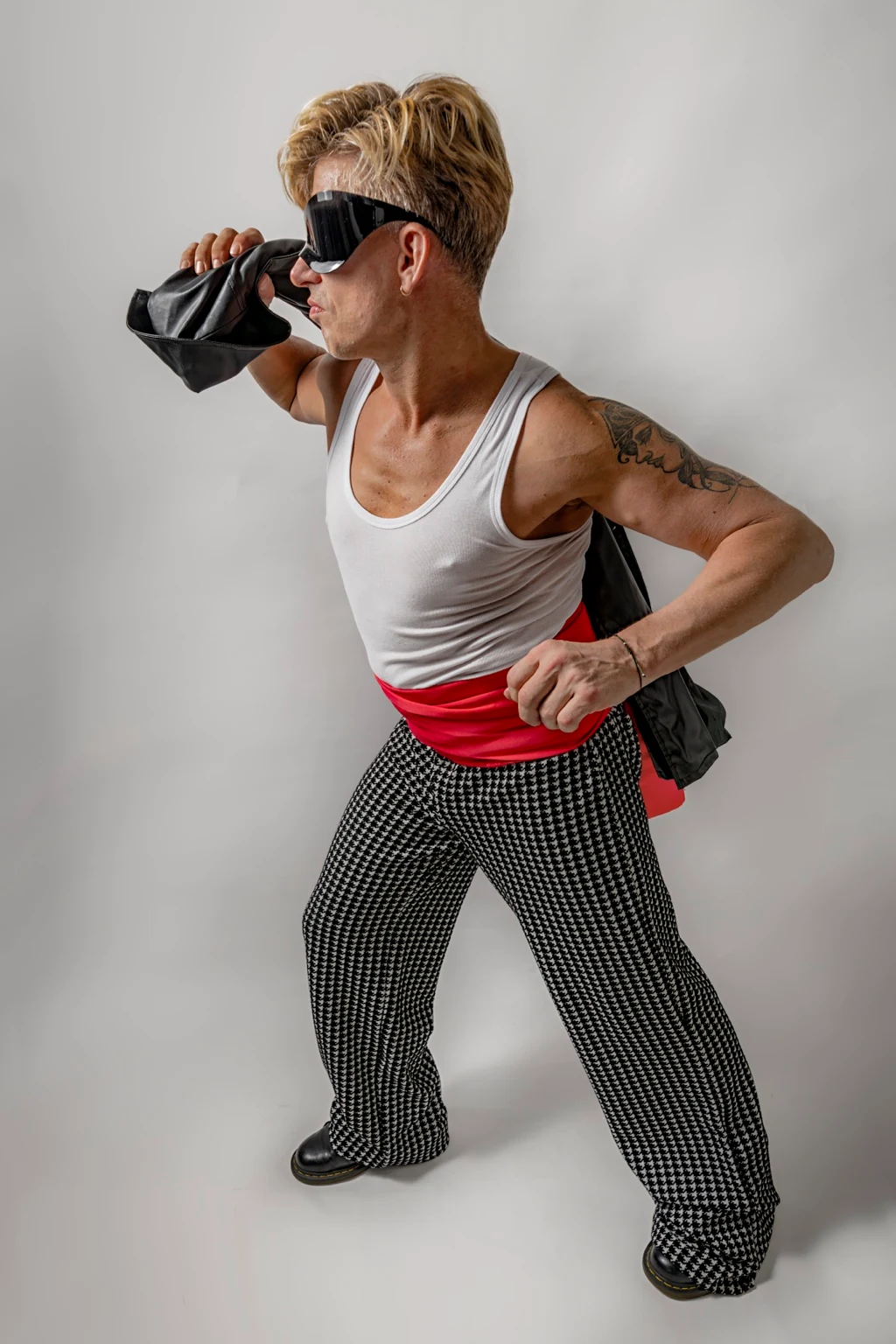 Self-portrait in dark goggles, a white vest, red sash and patterned trousers, holding black fabric at the shoulder.