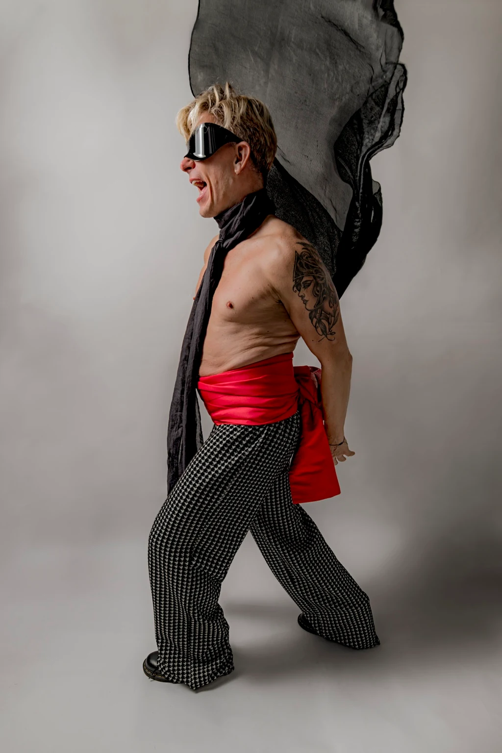Self-portrait in dark goggles, a red sash and patterned trousers, mouth open and a black scarf billowing overhead.